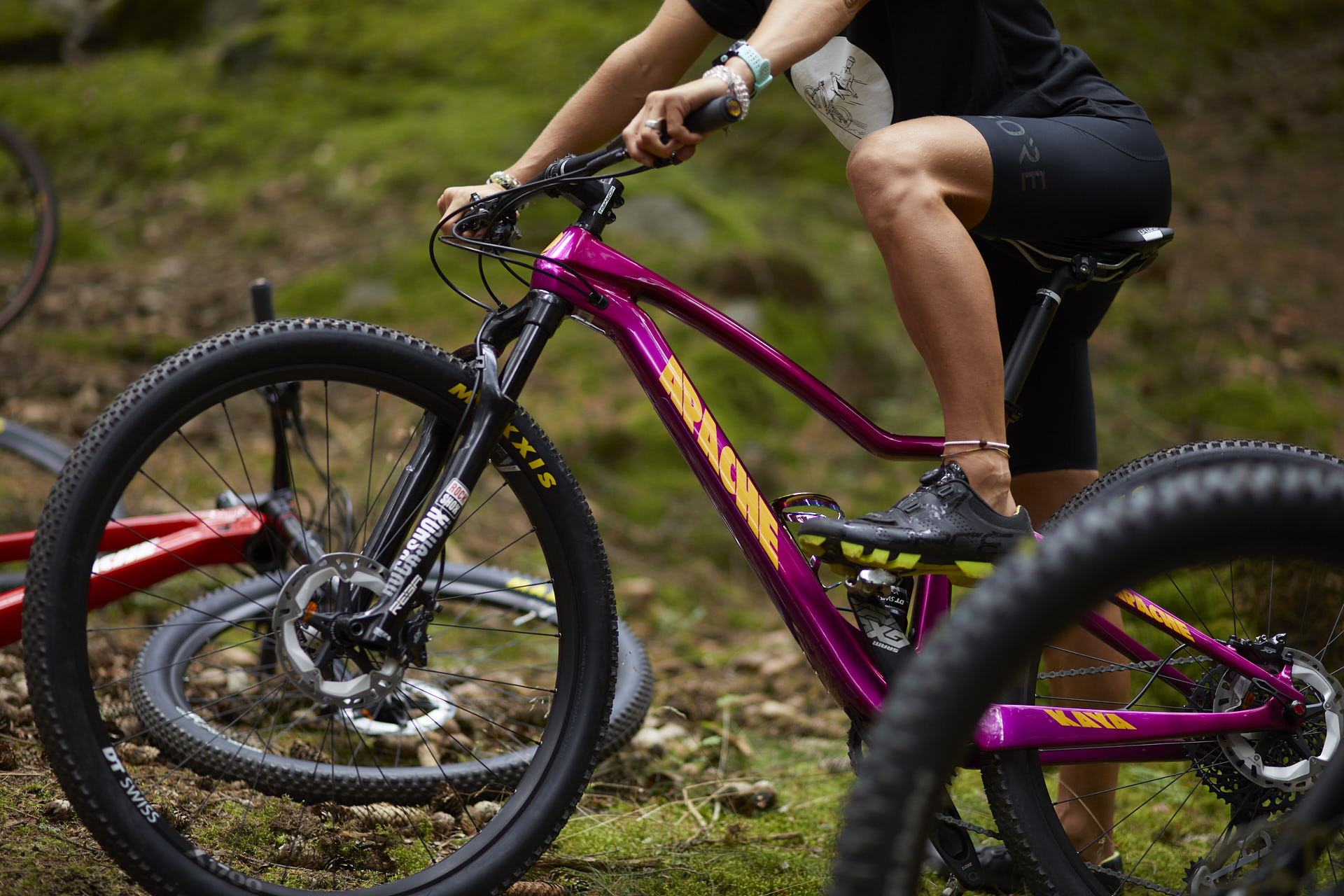 Close-up of a vibrant purple Apache mountain bike ride through a lush green forest, emphasizing the adventurous spirit of Apache's new visual identity. The bike's bold yellow Apache logo uses the slanted Speed Grotesk typeface, reflecting the rugged 'Mountains' category for challenging terrains. The sturdy Maxxis tires and RockShox fork add to the bike's performance-driven aesthetic, aligning with Apache Bikes' emphasis on durability and expressive typography.