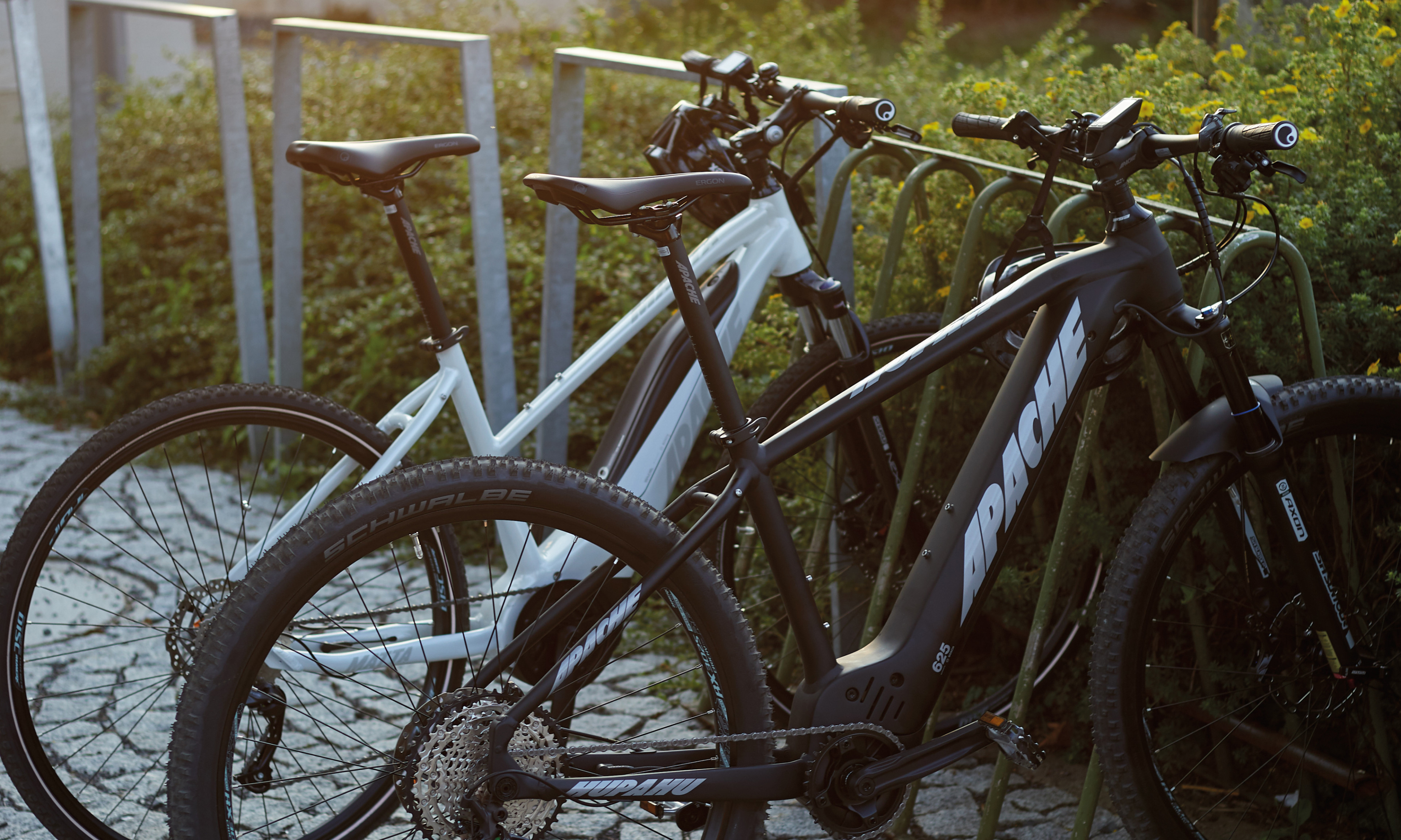 Two Apache Bikes, one in matte black and the other in white, are parked side by side on a bike rack. The matte black bike features Apache branding in bold white typography, reflecting the brand's expressive typography identity with robust design elements. These electric bikes, captured in soft natural lighting, embody the minimalist aesthetic and solid Speed Grotesk typeface introduced in Apache's new visual identity, highlighting the rugged yet modern design suitable for diverse terrains.