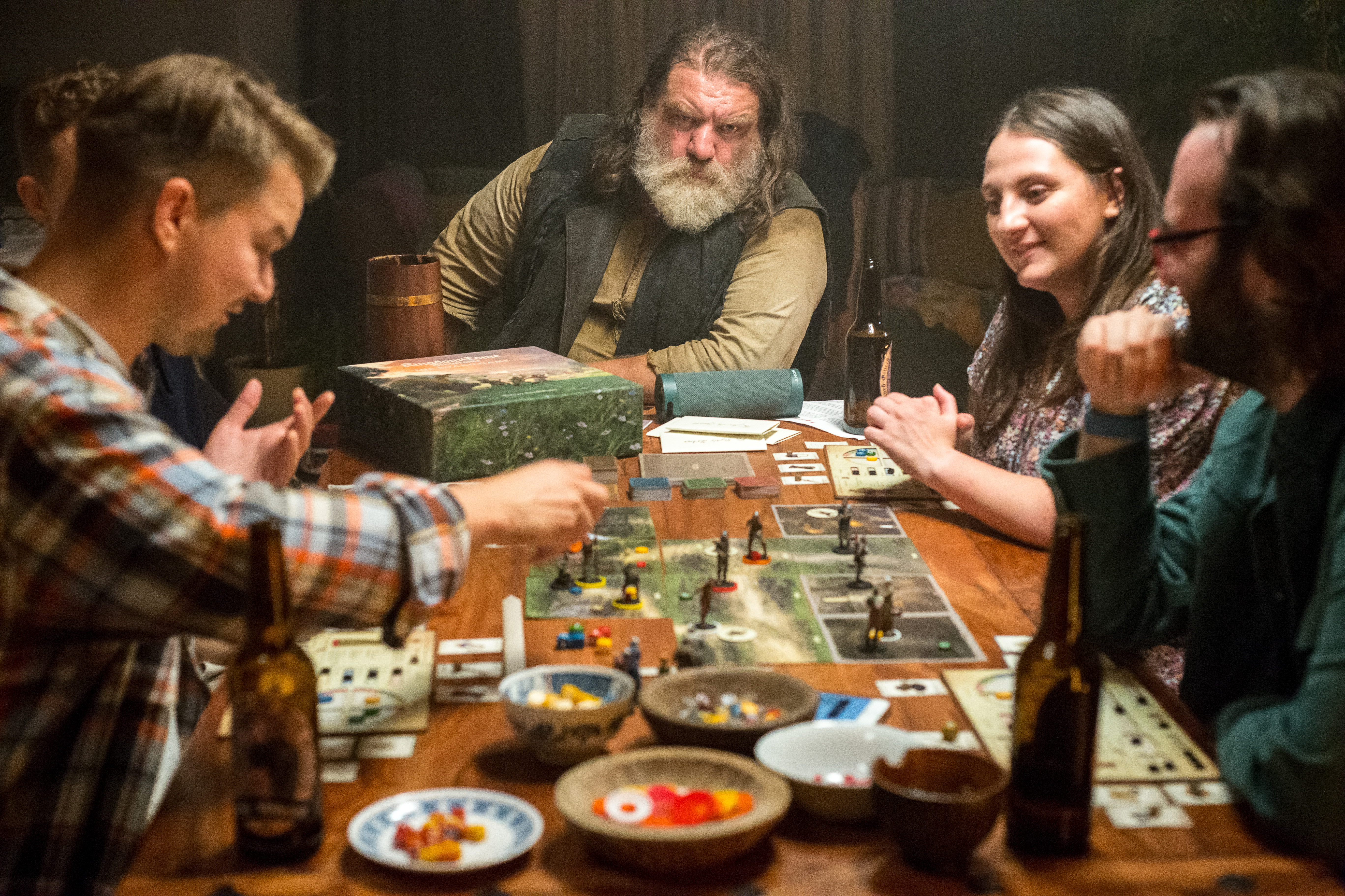 A group of players engaged in Kingdom Come: Deliverance board game gathered around a table with detailed game components and a mobile app, combining physical and digital elements to deepen story possibilities and interactive gameplay.