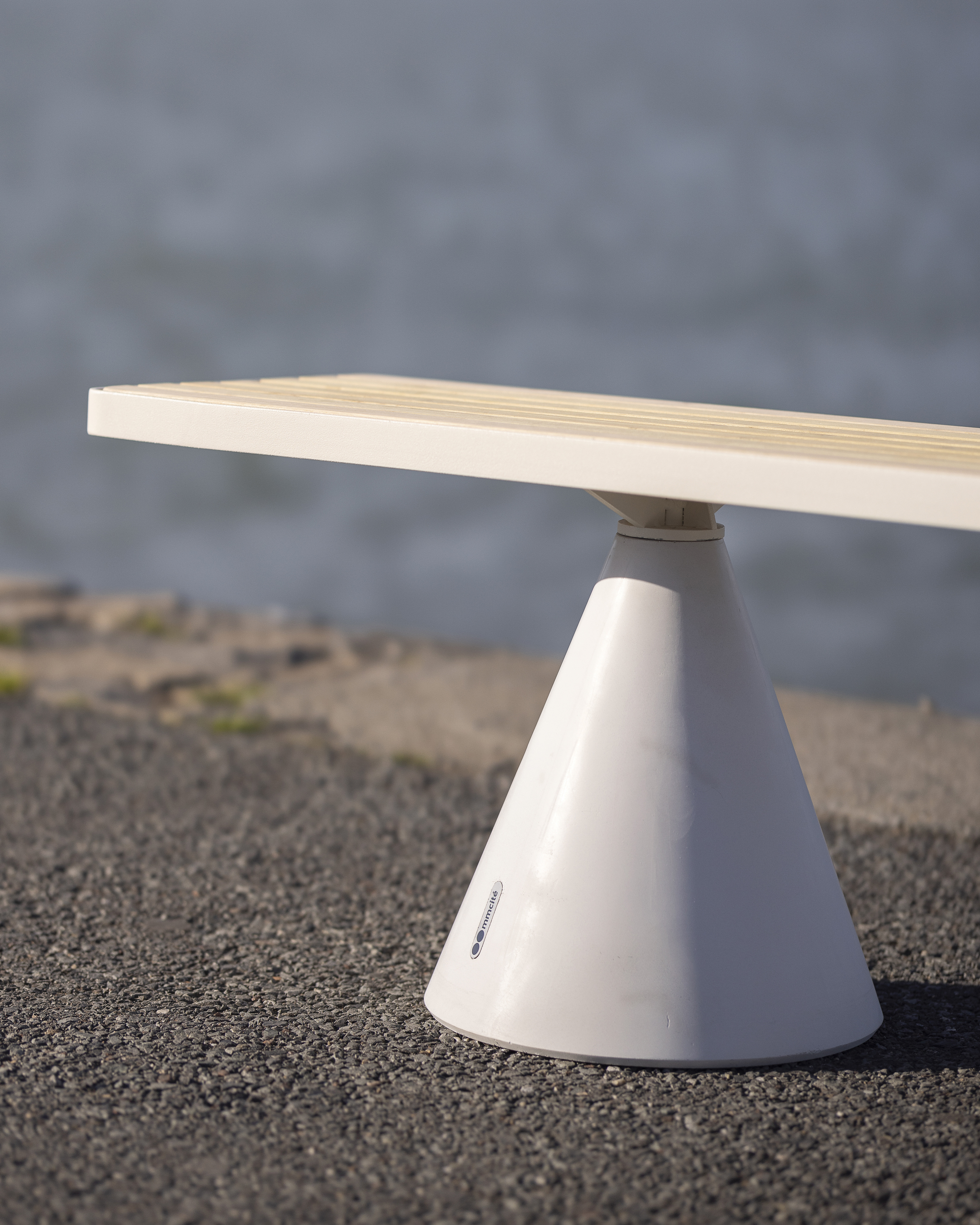Close-up on a Logotype of a minimalist bench by mmcité, featuring a white conical base with a flat wooden seat, reflecting the brand's rational design update and focus on functional aesthetics.