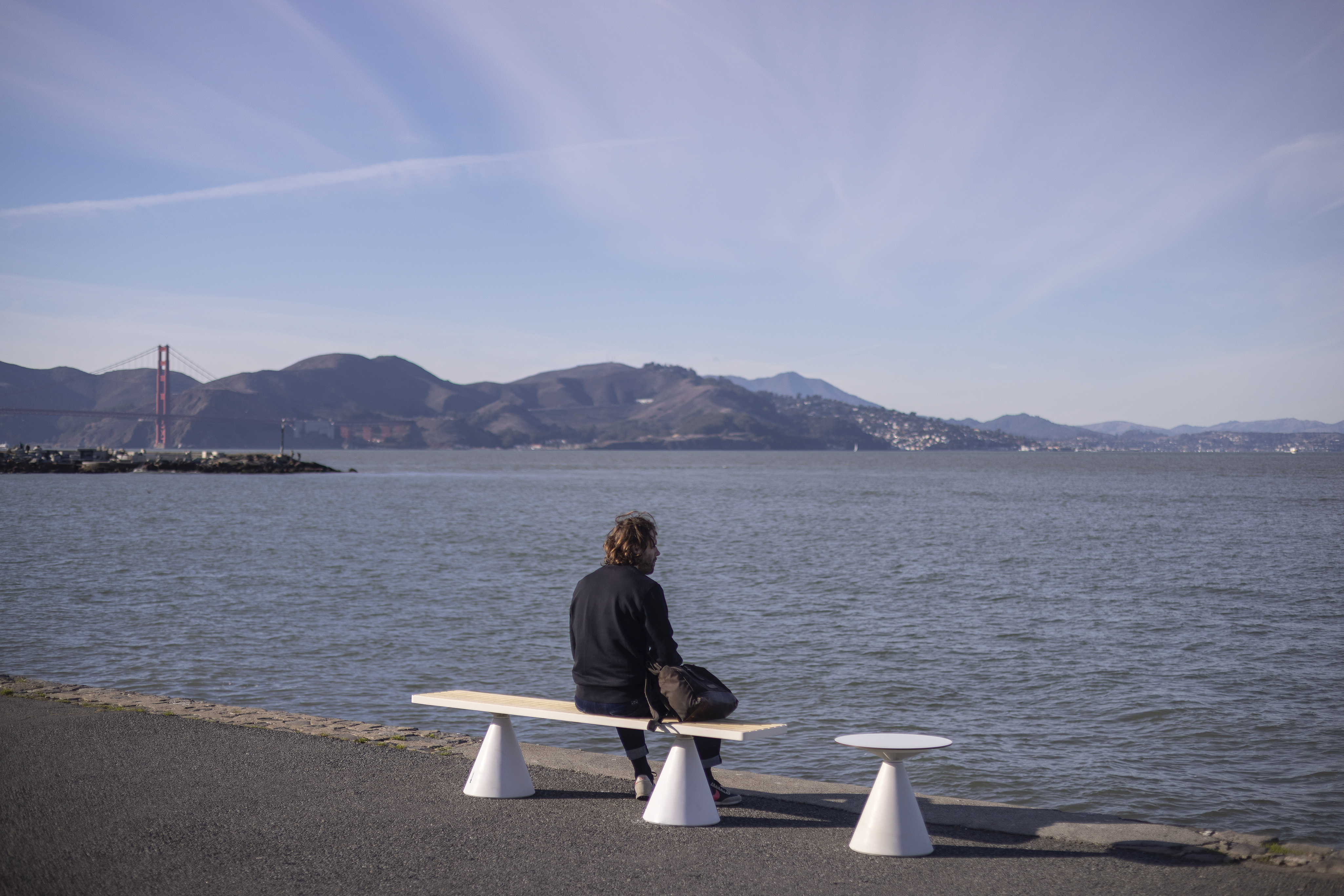 Zoom out the same photo as the first picture where Person is sitting on a mmcité bench along a waterfront, with the Golden Gate Bridge in the background, embodying mmcité's approach to blending rational design with natural landscapes in urban settings.