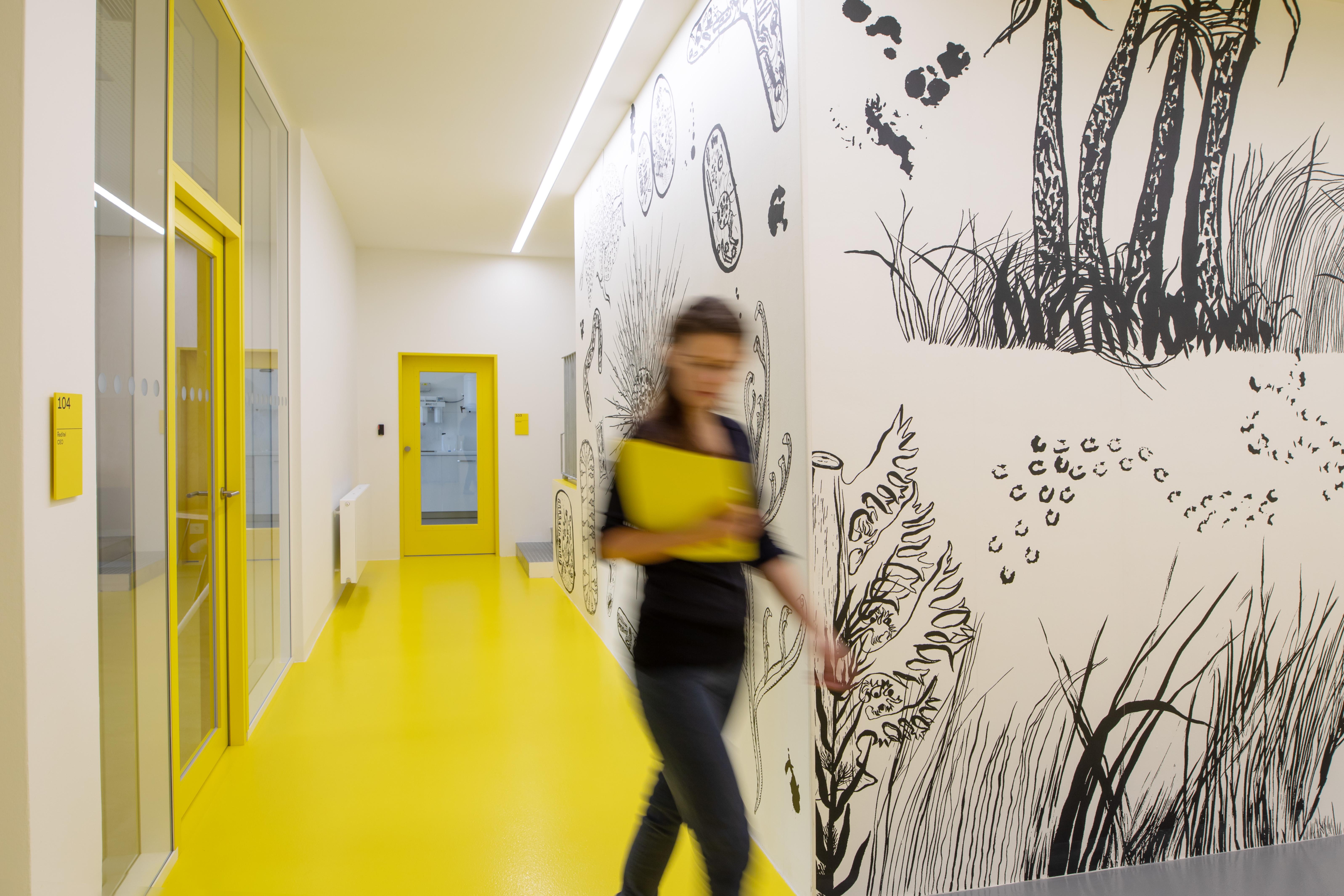 The interior hallway of Naturfyt Bio's factory has bright yellow flooring and door frames, complemented by black-and-white hand-drawn botanical illustrations on the walls, representing Naturfyt Bio's branding that integrates nature-inspired graphics with functional design.